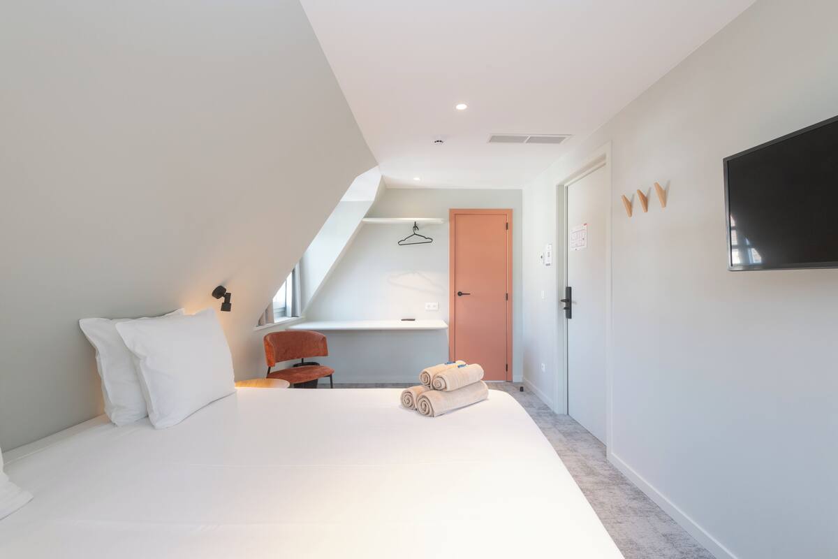 A modern hotel room with a large bed, a desk, a chair, and a TV mounted on the wall.