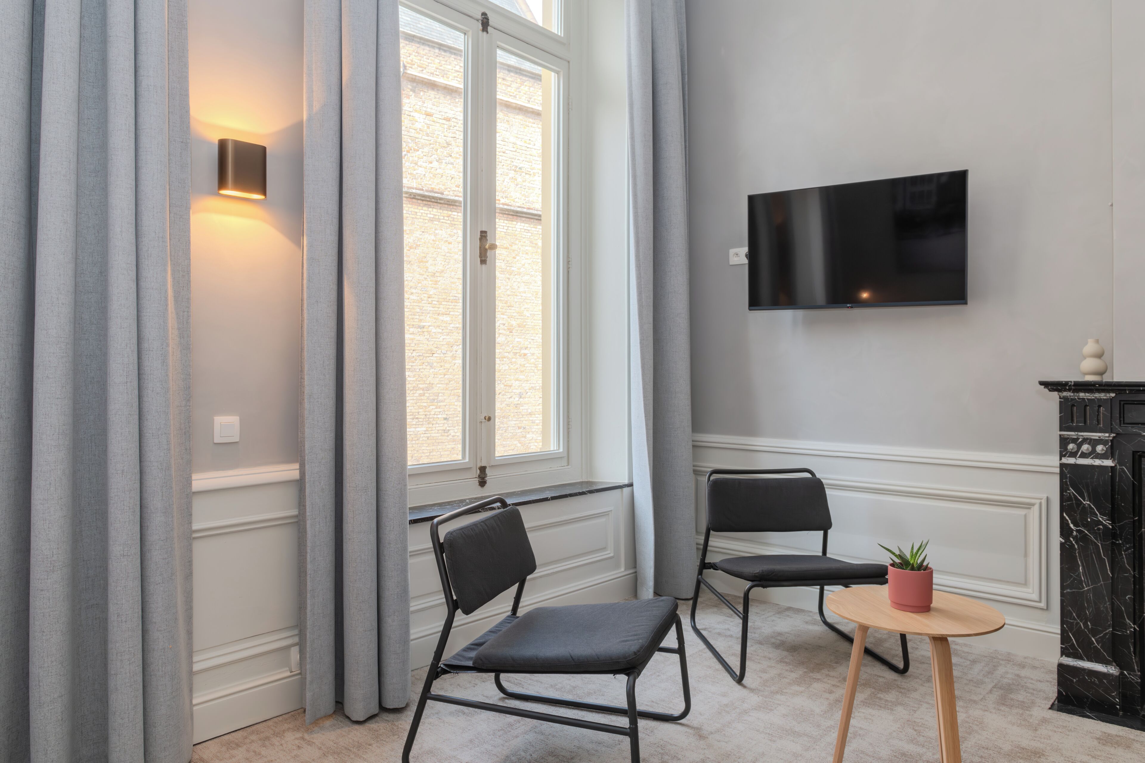 Una habitación con un ventanal grande, dos sillones, una mesita con una planta, un televisor montado en la pared y una chimenea.