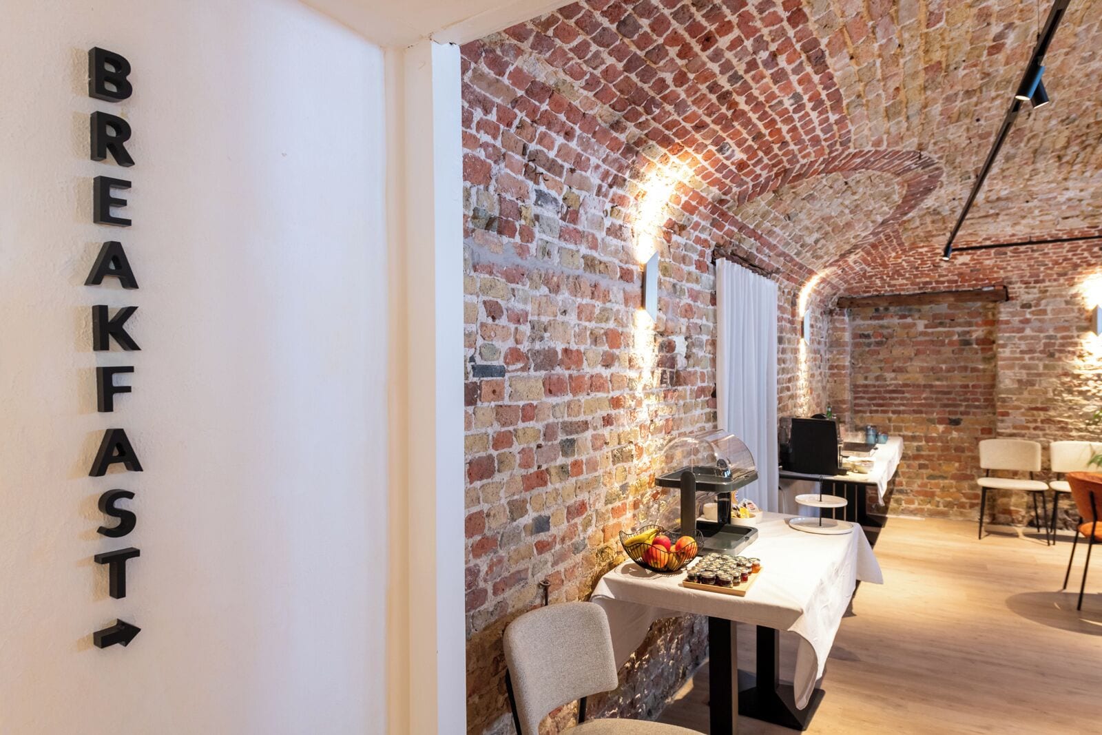 A breakfast area with exposed brick walls, a table with a fruit basket, and a sign that reads "BREAKFAST".