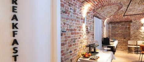 A breakfast area with exposed brick walls, a table with a fruit basket, and a sign that reads "BREAKFAST".