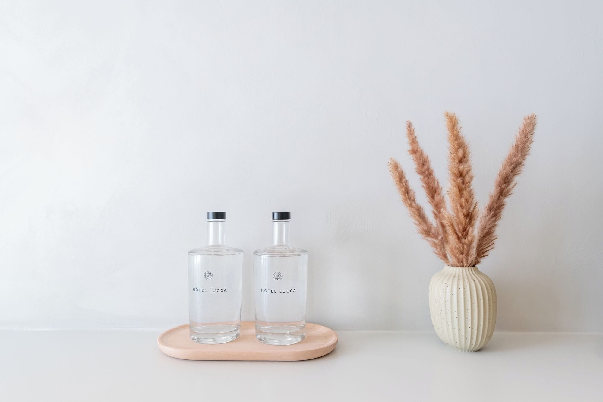 Dos botellas de vidrio transparente con tapas metálicas, identificadas con 'HOTEL LUCCA', sobre una bandeja de madera sobre un fondo liso.