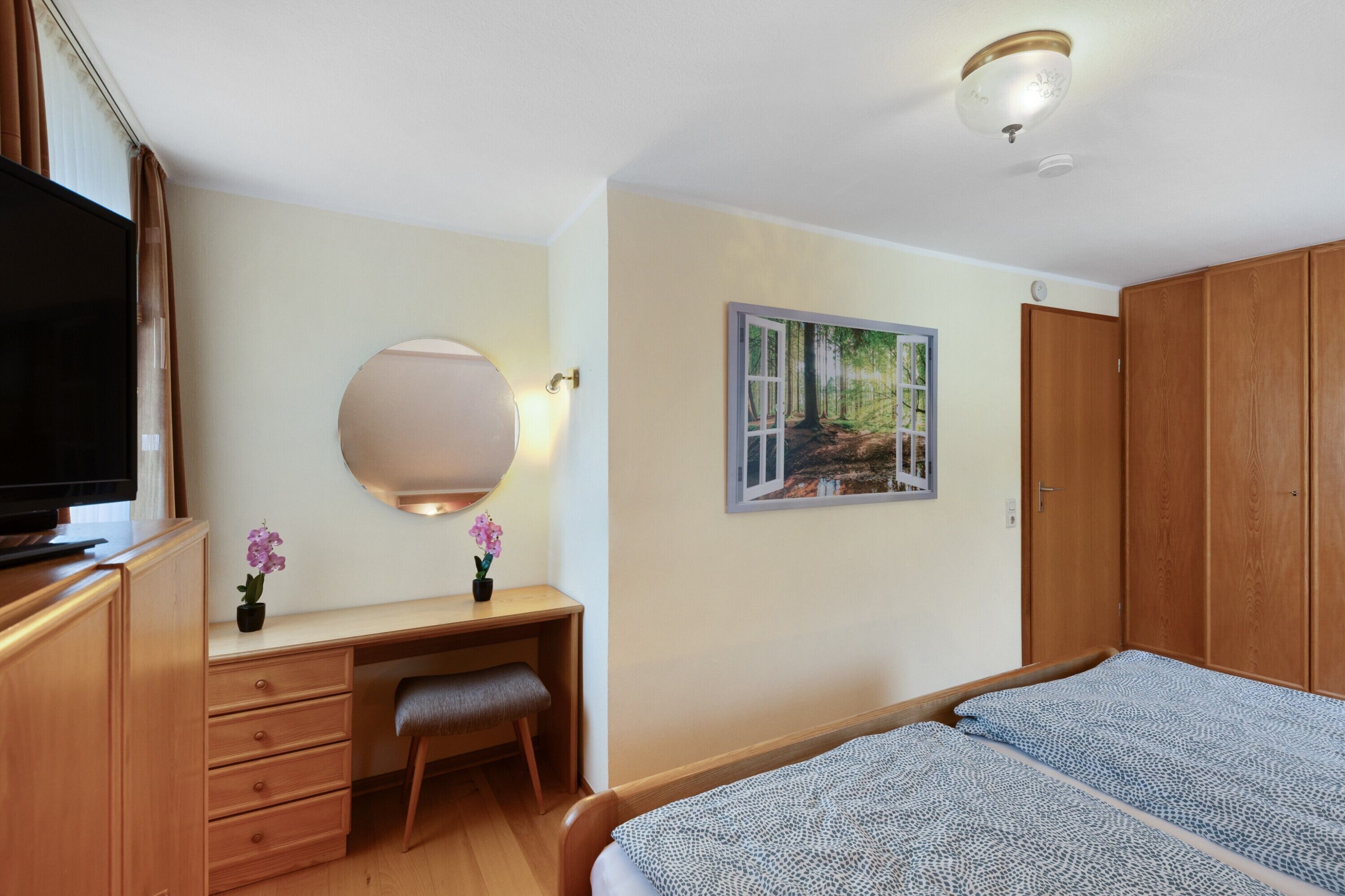 Ein Schlafzimmer mit Bett, Schreibtisch mit Schubladen, Fernseher, Spiegel und einem Bild an der Wand.
