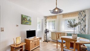 A living room with a flat-screen TV, wooden furniture, a fan, and a window with curtains. - Apartment '2 Bergblick, 60sqm' with Mountain View, Shared Garden and Wi-Fi (Lennestadt)