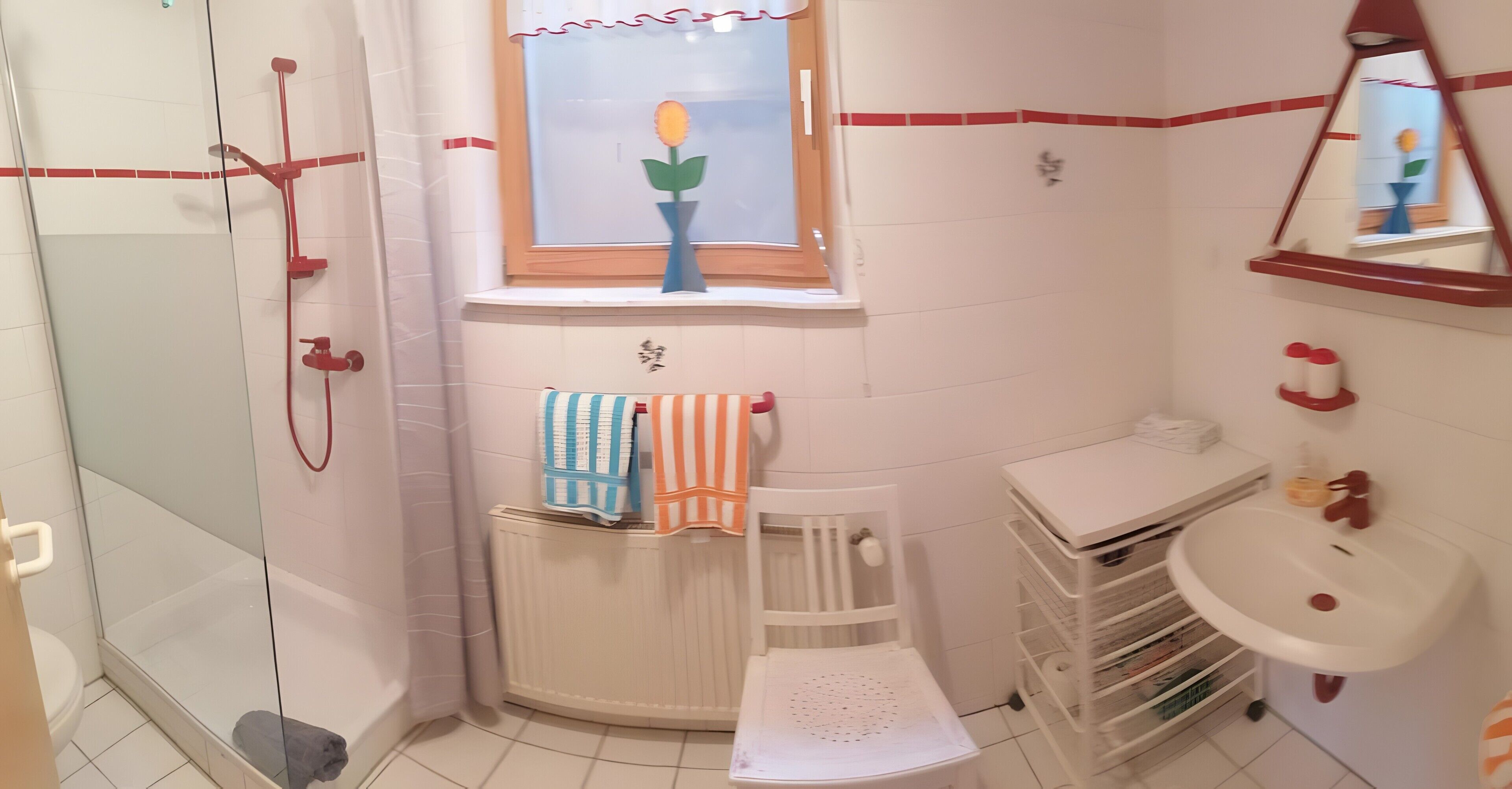 A bathroom with a glass shower enclosure, a white radiator, a window with a flower vase, a white sink, and a white chair.