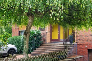 Un bâtiment en briques avec une porte jaune, un escalier menant à l’entrée et un arbre qui offre de l’ombre.