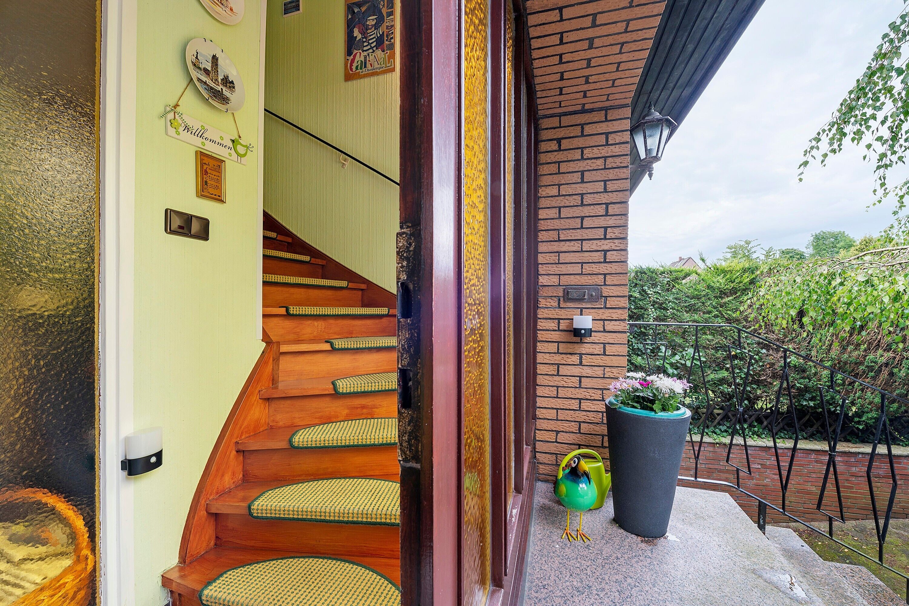 Eine Holztreppe führt zu einer Ziegelhausfassade, mit einem Blumentopf und einem Willkommensschild.