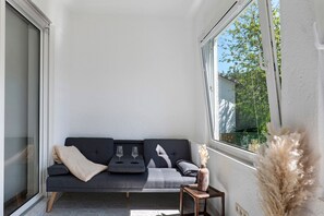 Un interior moderno con un sofá gris, dos copas de vino, un jarrón y una ventana con vista a zonas verdes.