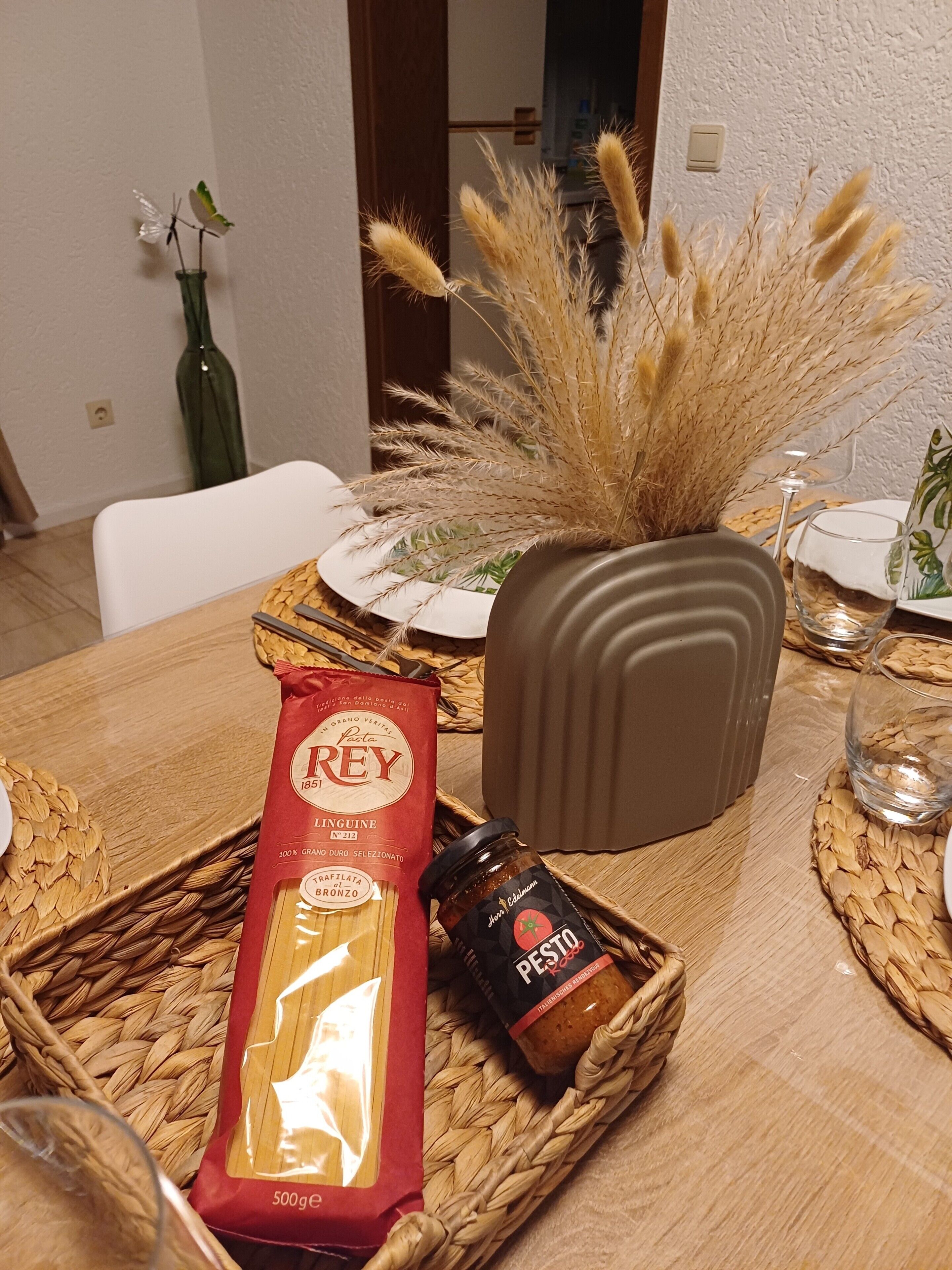 A dining table with a woven placemat, a glass of water, a jar of pesto, a packet of pasta, and a decorative vase with dried plants.