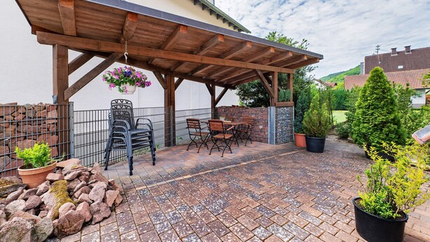 Een overdekt terras met een houten pergola, buitenmeubilair, plantenpotten en een pad van bakstenen.