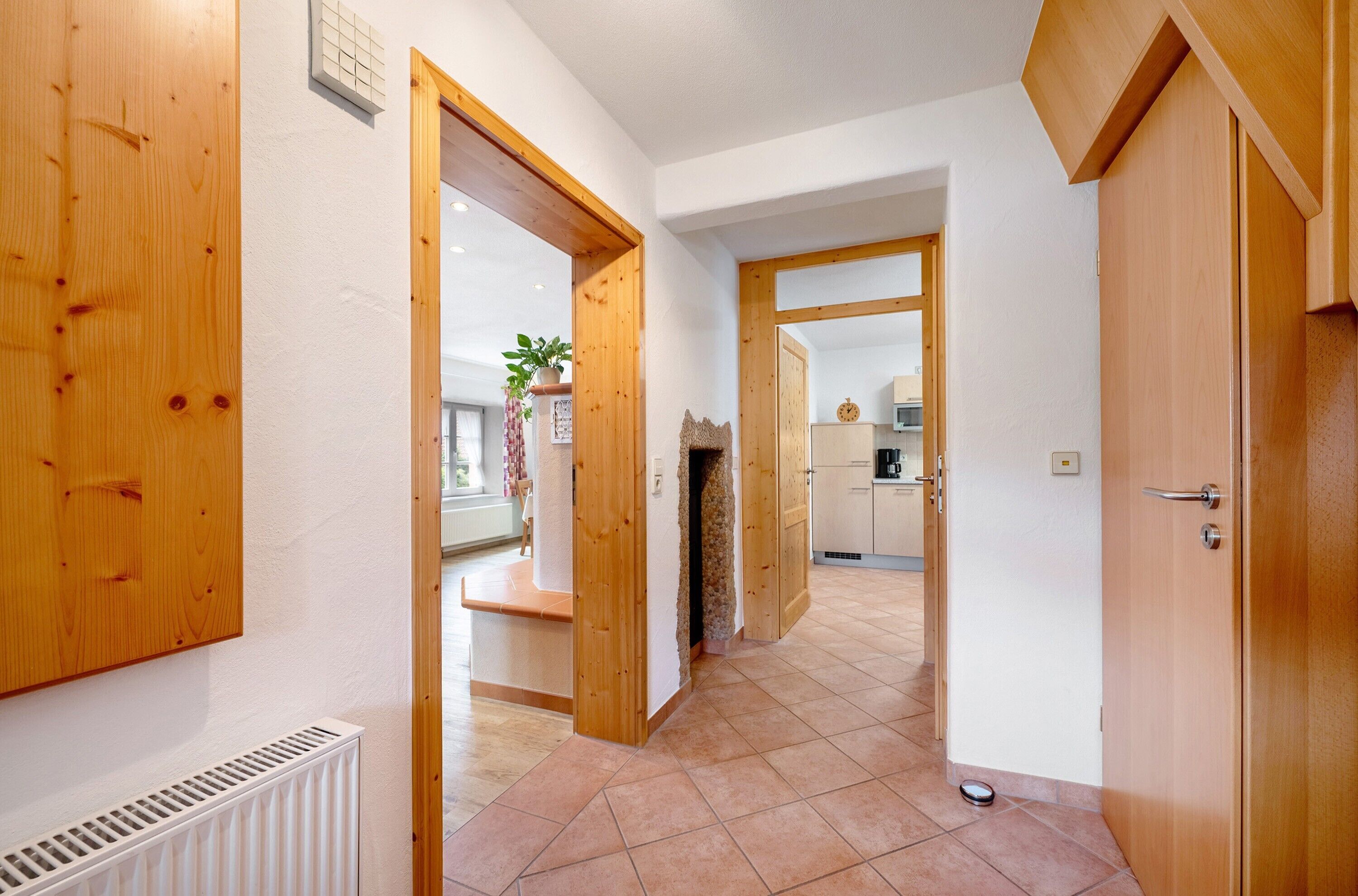 Un couloir avec des portes en bois, un radiateur et un sol carrelé qui mène à un espace cuisine.