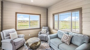 A room with a view of a grassy field, a gray sofa, a gray armchair, and a patterned ottoman. - Ayu Ranch | Panoramic views | Hot tub (Cheyenne)