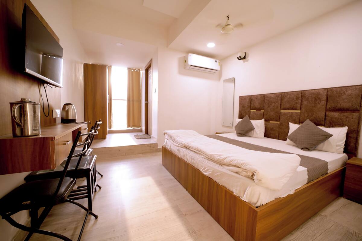 A bedroom with a wooden bed, a TV mounted on the wall, a desk with a kettle, and a chair.