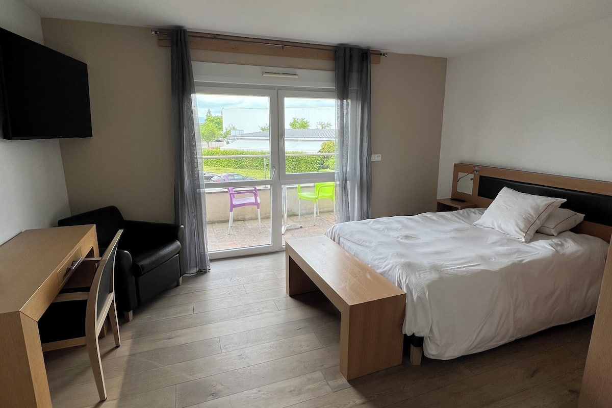 Une chambre d’hôtel avec un lit, un bureau et une chaise. Elle dispose d’un balcon avec vue sur l’extérieur.