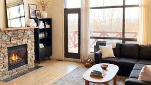 A living room with a stone fireplace, a black sofa, a coffee table, and a large window with curtains. - Ski-In/Out Mountain Escape with Hot Tub (Kimberley)