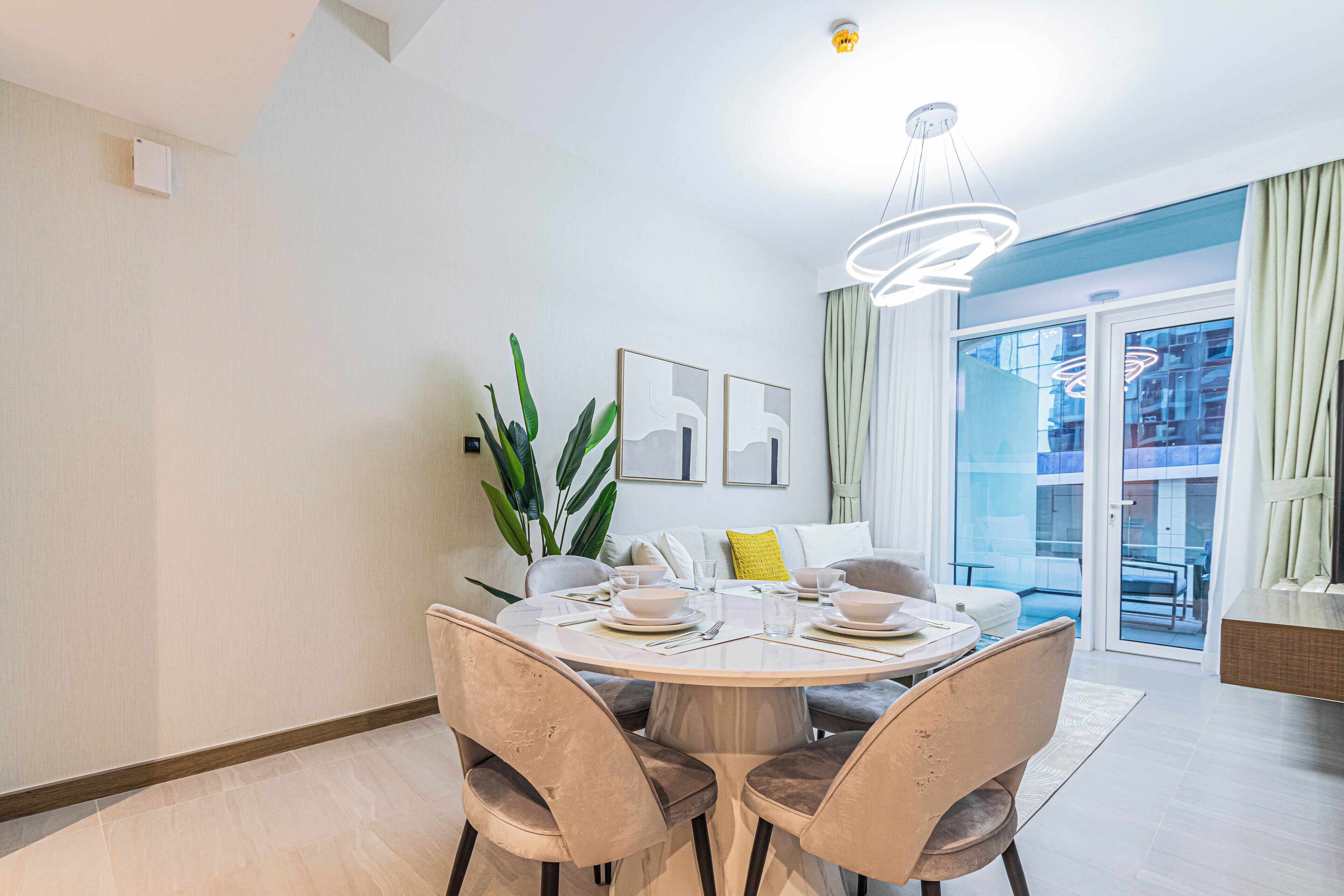 Un comedor con una mesa redonda dispuesta para cuatro personas, rodeada de sillas. Hay una planta a la izquierda de la mesa y un sofá con un cojín amarillo al fondo.