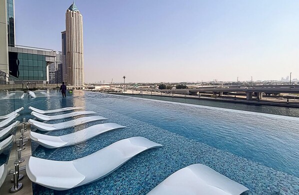 Piscina con sillas de descanso blancas y modernas con forma de ola, y vista a un paisaje urbano con edificios altos.