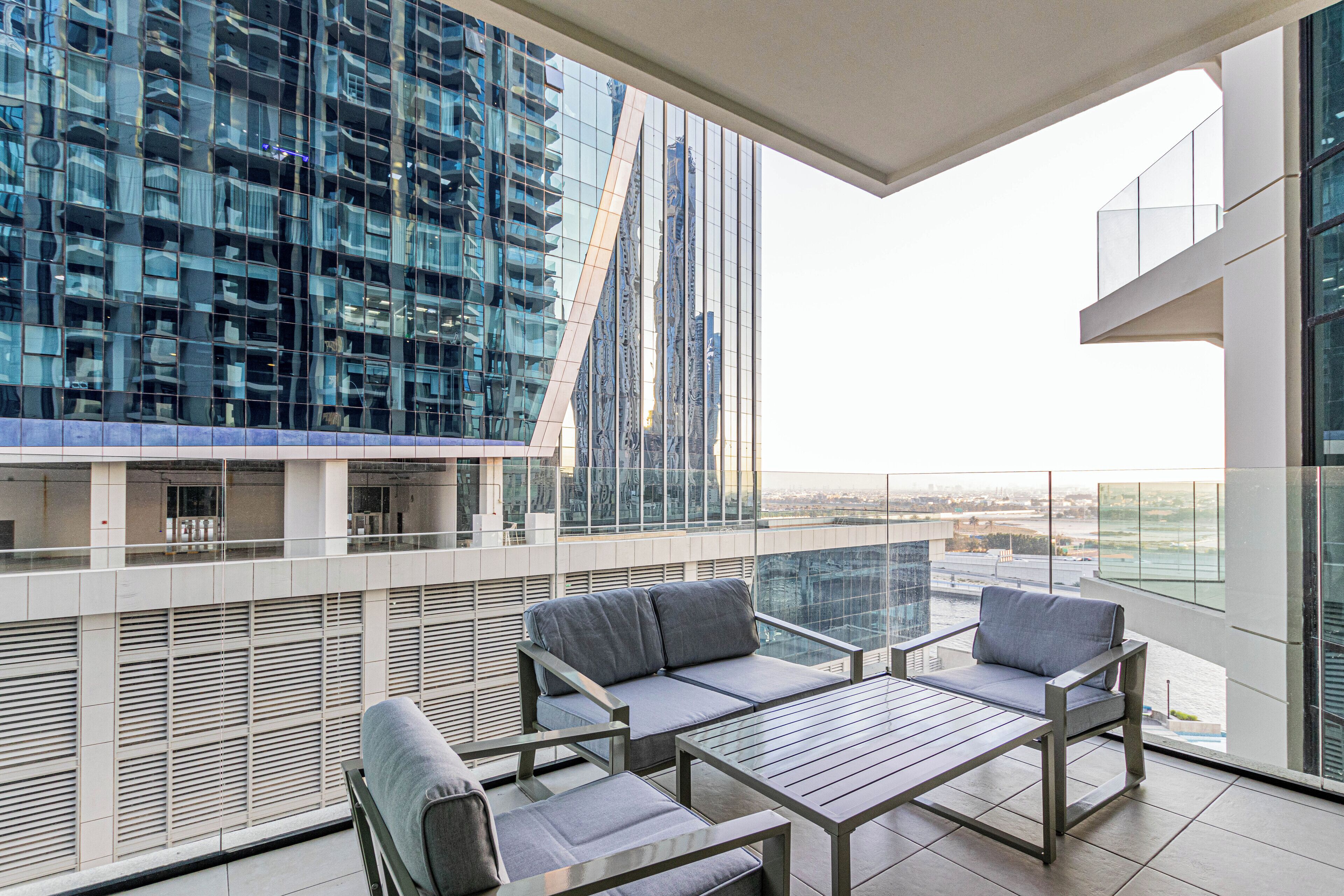 A modern balcony with a glass railing, a sofa, and a table, overlooking a cityscape with high-rise buildings.