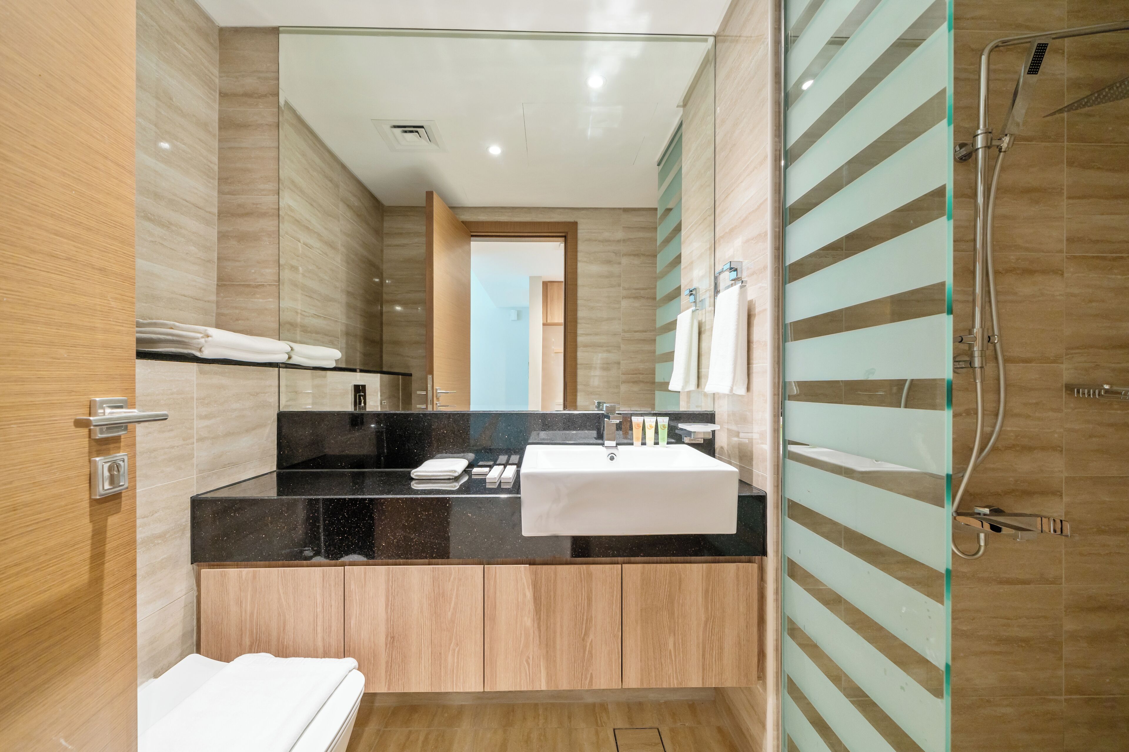 A modern bathroom with a black countertop, wooden cabinets, a glass shower enclosure, and a white sink.