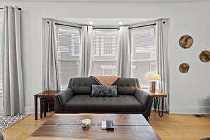 Un salon avec un canapé foncé, une table basse, une lampe et une fenêtre avec des stores.