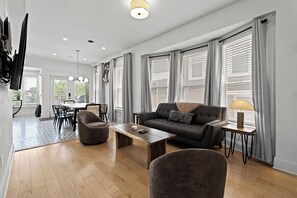 Ein modernes Wohnzimmer mit einer Couch, Sesseln, einem Couchtisch und einem an der Wand montierten Fernseher.