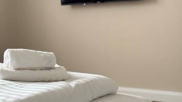 Una cama bien hecha con sábanas blancas y una toalla doblada encima, un televisor de pantalla plana montado en la pared y suelo de madera.