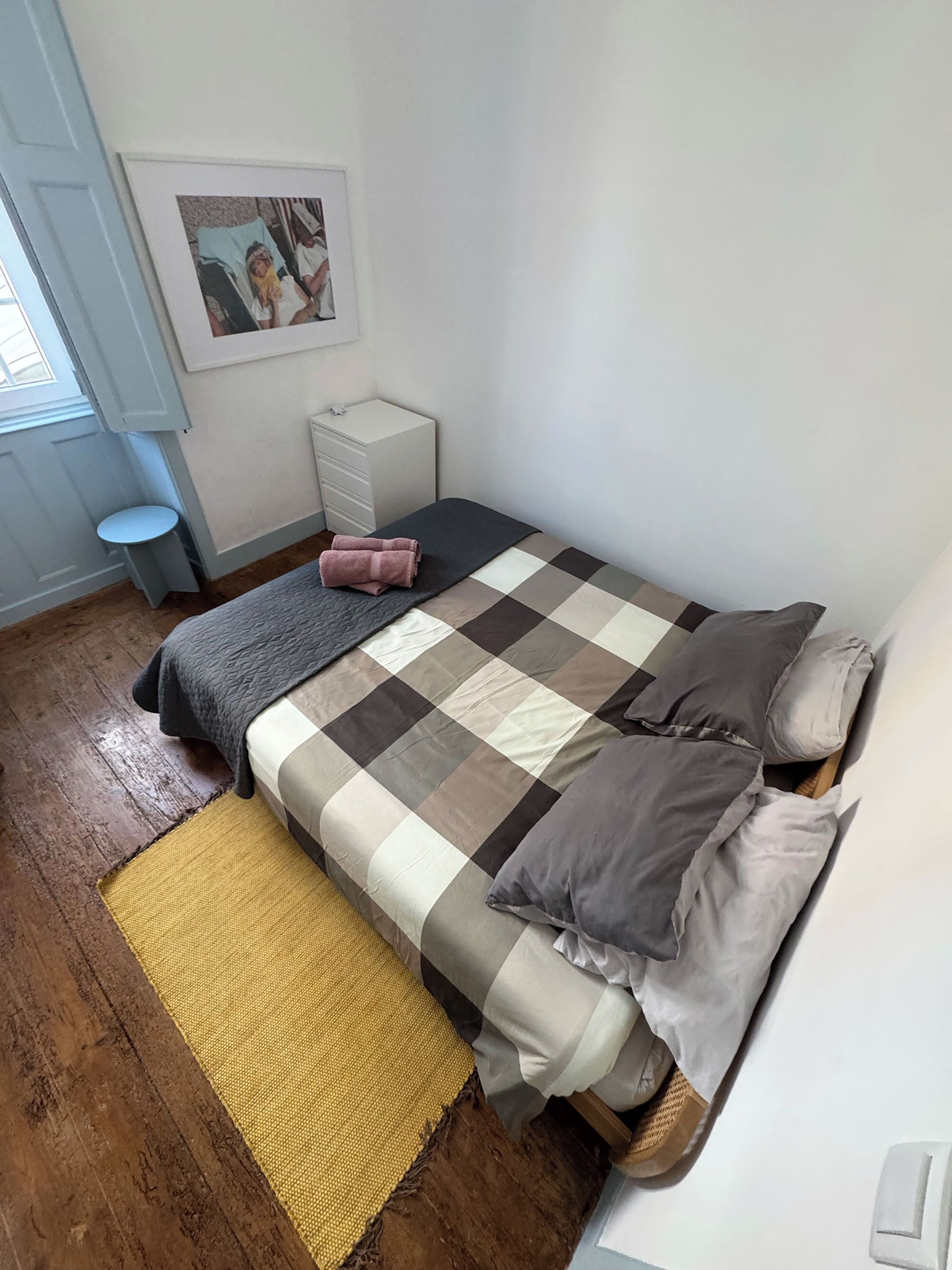 Ein Schlafzimmer mit einem Bett, einem Nachttisch, einem kleinen Hocker, einem Bild an der Wand und einem Fenster.