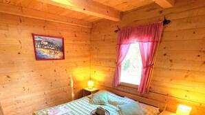 Egy hangulatos, fából készült kabin, amelyben egy ágy, egy éjjeliszemélyzet, egy rózsaszín függönnyel ellátott ablak és egy képkeretben elhelyezett kép található a falon.