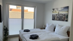Ein Schlafzimmer mit großem Fenster, einem Bett mit weißer Bettwäsche und einem Stadtbild an der Wand.