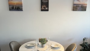 Ein Essbereich mit einem runden Tisch, gedeckt für vier Personen, umgeben von grauen, gepolsterten Stühlen. Über dem Tisch hängen zwei gerahmte Kunstwerke, eines zeigt eine Strandansicht, das andere eine Kaffeetasse mit Text. Rechts ist eine kleine Topfpflanze zu sehen.