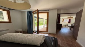 Une chambre à coucher avec un lit, un petit tabouret, une fenêtre avec des rideaux et une porte ouverte donnant sur un balcon.