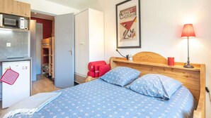 A bedroom with a bed, a wooden headboard, a red chair, a small table, a lamp, a microwave, a refrigerator, and a poster on the wall. - Studio in Chamrousse near Ski Resort (Chamrousse)