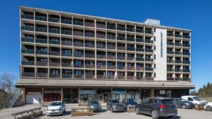 A multi-story hotel with a central entrance, multiple balconies, and a parking area with several cars. - Studio in Chamrousse near Ski Resort (Chamrousse)