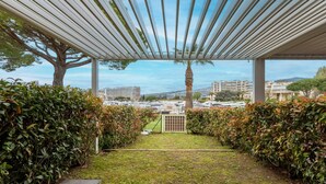 Ein überdachter Außenbereich mit Blick auf eine Stadtlandschaft, mit einer Pergola, einer Palme und ordentlich geschnittenen Hecken.