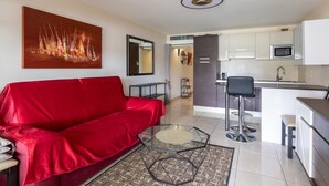 Ein modernes Wohnzimmer mit einem roten Sofa, einem Glastisch und einem großen abstrakten Gemälde. Im Hintergrund ist eine Küche mit weißen Schränken und einer Mikrowelle zu sehen.