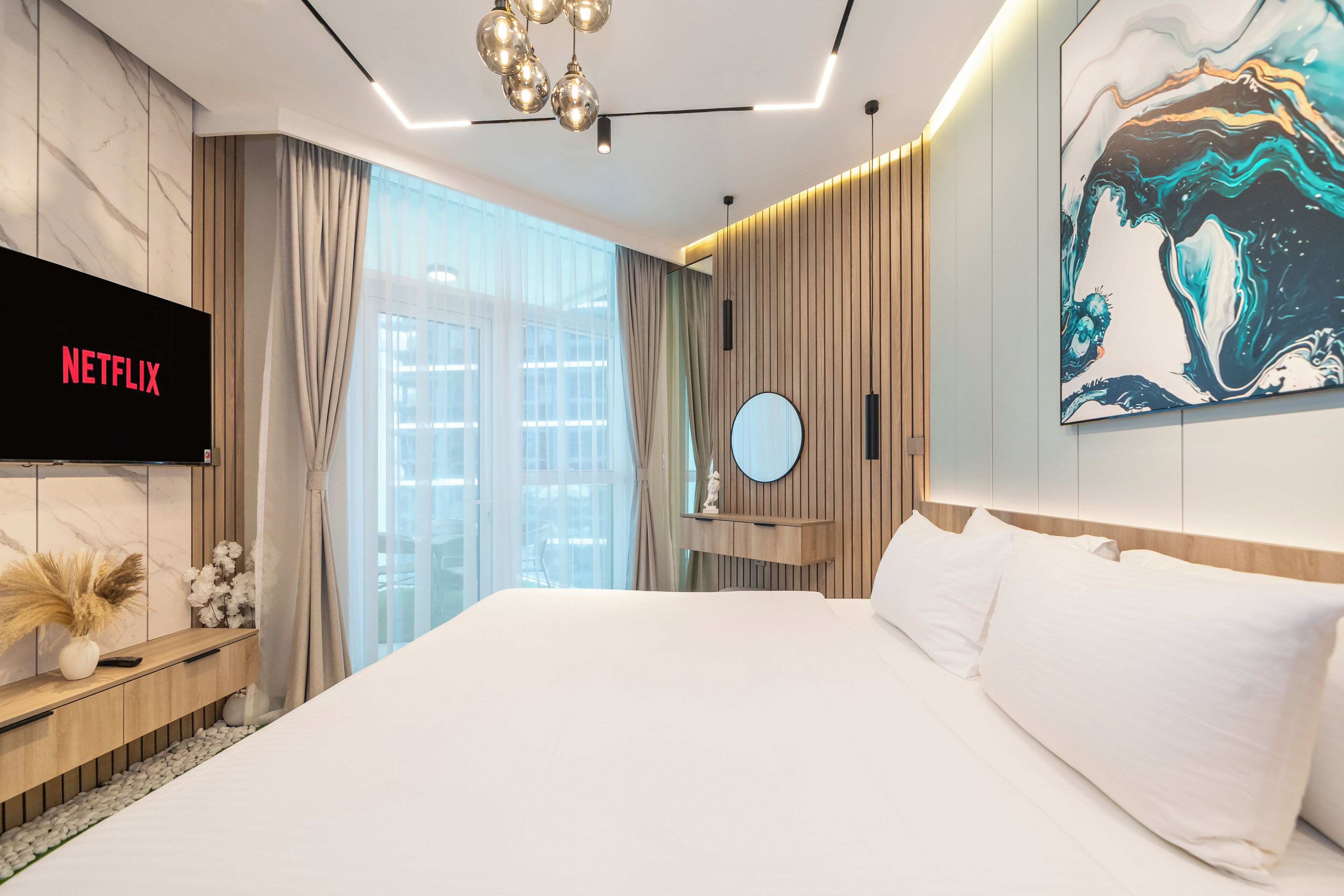 Quarto de hotel moderno com uma cama grande, uma TV fixada exibindo 'NETFLIX', uma janela ampla com cortinas e uma obra de arte contemporânea na parede.