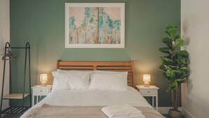 A neatly made bed with white linens, two bedside tables with lamps, a wooden headboard, a wall-mounted painting, and a potted plant. - Modern 3BR Home with Stunning Garden | Banbury Stay (Banbury)