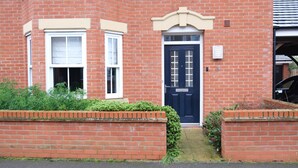 Une maison en briques rouges, avec une porte d’entrée bleu foncé, des fenêtres à cadre blanc et un petit jardin agrémenté de plantes vertes.
