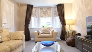 Un salon avec un canapé beige, un fauteuil beige agrémenté de coussins décoratifs, une table basse en verre bleu, un téléviseur et une lampe.