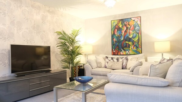 Salón con televisor de pantalla plana, una mesa de centro de vidrio con un tazón azul, un sofá con cojines estampados, un cuadro abstracto grande y una planta en maceta.