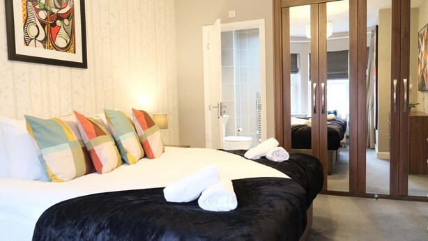 Une chambre d’hôtel avec un grand lit, des oreillers colorés, une tête de lit noire, un miroir et une porte coulissante donnant sur une salle de bain.
