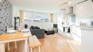 En moderne lejlighed med spiseplads, sofa, TV og et køkken med hvide skabe og et stort vindue.