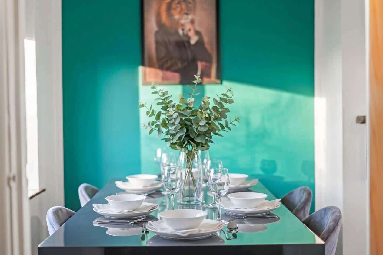 A dining table set with plates, glasses, and a vase of greenery, against a teal wall with a framed picture.