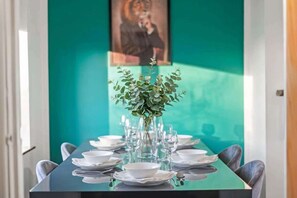 A dining table set with plates, glasses, and a vase of greenery, against a teal wall with a framed picture. - Modern 3BED Home in Rugby - Perfect for Groups (Rugby)