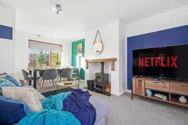 A living room with a fireplace, a dining area, and a large TV screen displaying 'NETFLIX'.
