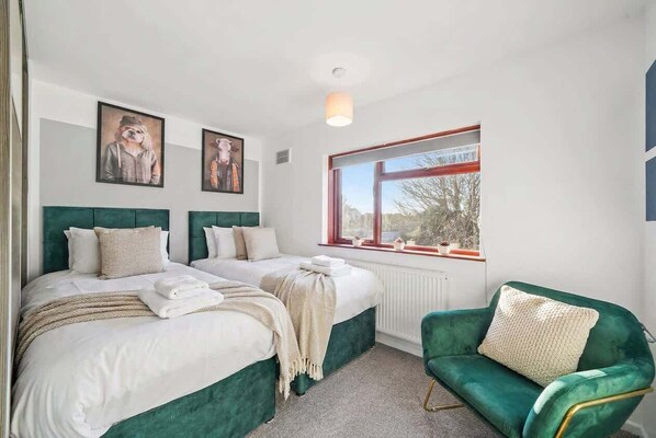 A hotel room with two beds, a green armchair, and a window with a view.