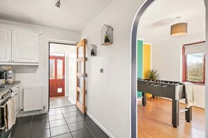 A kitchen with white cabinets, a stove, and a tiled floor. There is a foosball table in a room with a wooden floor and a window.