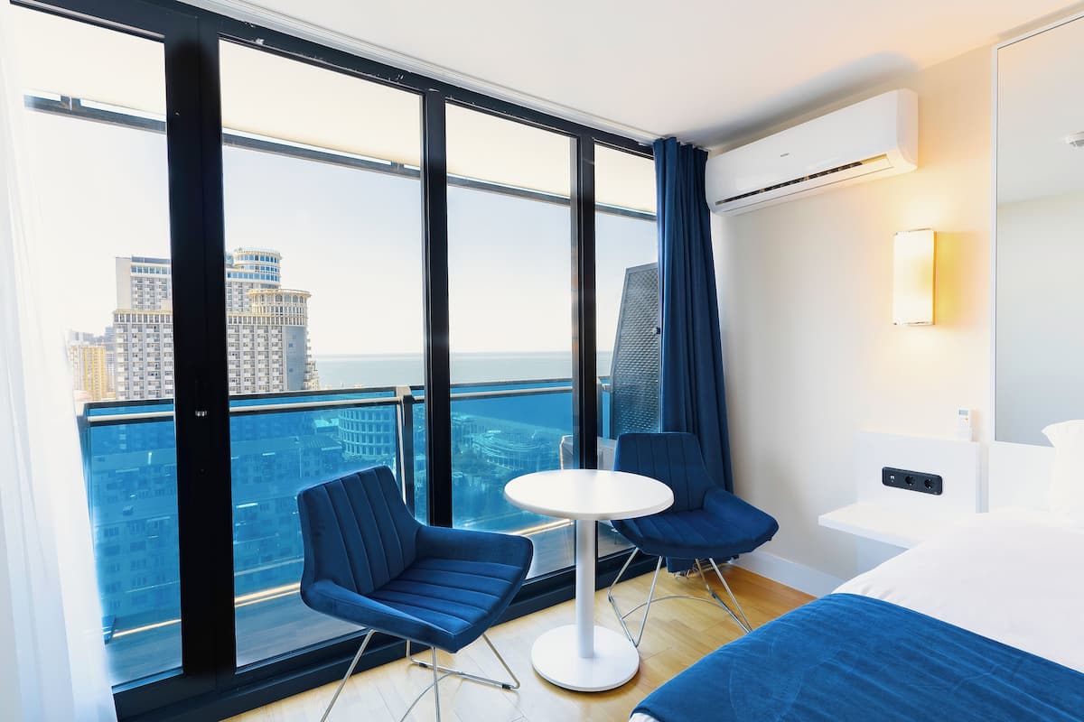 Une chambre d’hôtel moderne avec une grande fenêtre, une table ronde, deux chaises bleues, un lit et une table de chevet avec une lampe.