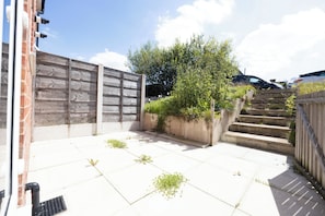 A paved patio area with a wooden fence, a concrete planter with plants, and a staircase leading to a higher level.