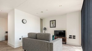 A modern living room with a grey sofa, a flat-screen TV, and a wall-mounted clock. - Elegant 1BR – Sheffield City Centre (South Yorkshire)