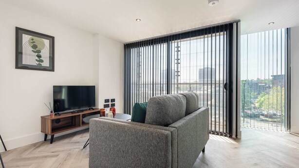 A modern living room with a sofa, a TV on a wooden stand, and a large window with blinds. - Elegant 1BR – Sheffield City Centre (South Yorkshire)